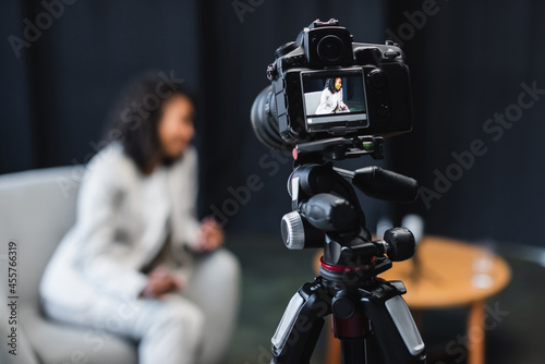 modern digital camera on tripod with african american journalist sitting in armchair on screen