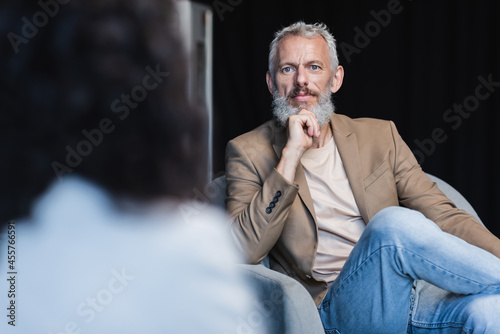 confident businessman sitting in armchair near blurred african american journalist during interview