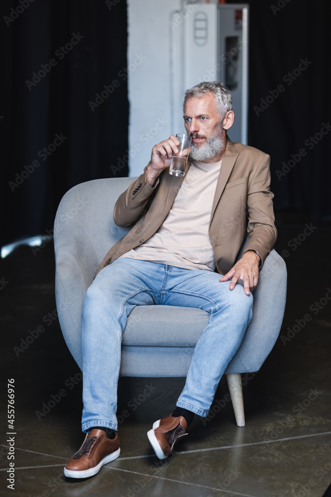 bearded businessman holding glass of water and sitting in grey armchair during interview bearded businessman holding glass of water and sitting in grey armchair during interview