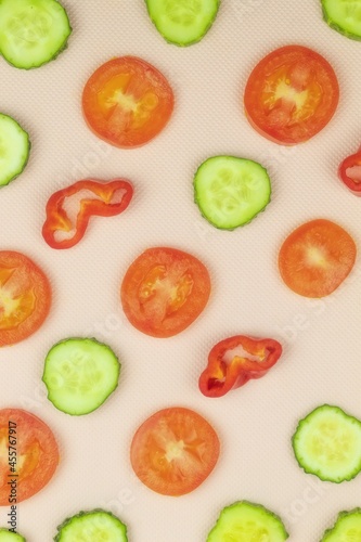 On the pink background of the vertical photo are round slices of green cucumber, red tomato and sweet pepper