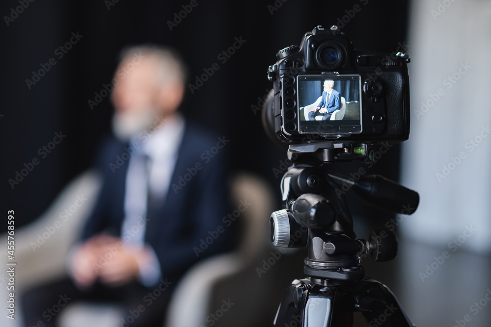 digital camera with businessman in suit sitting in grey armchair during interview on screen