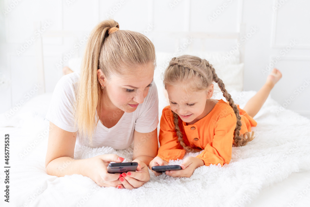 mom and daughter with mobile phones are lying on the bed at home and taking selfies or talking via video link mom and daughter with mobile phones are lying on the bed at home and taking selfies or talking via video link