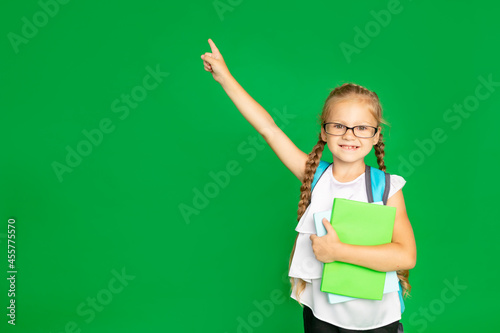 a schoolgirl on an isolated green background with books, textbooks and a backpack in glasses points her finger to the side, space for text