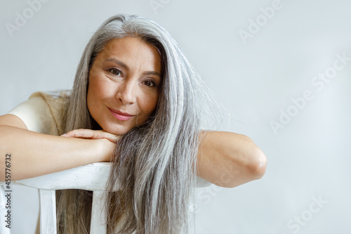 Cheerful middle aged Asian lady with flowing hoary hair sits backwards on chair on light background in studio. Mature beauty lifestyle