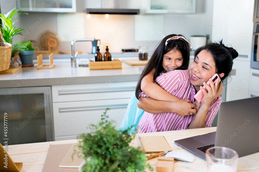 Mother working at home with her daughter hugging her 