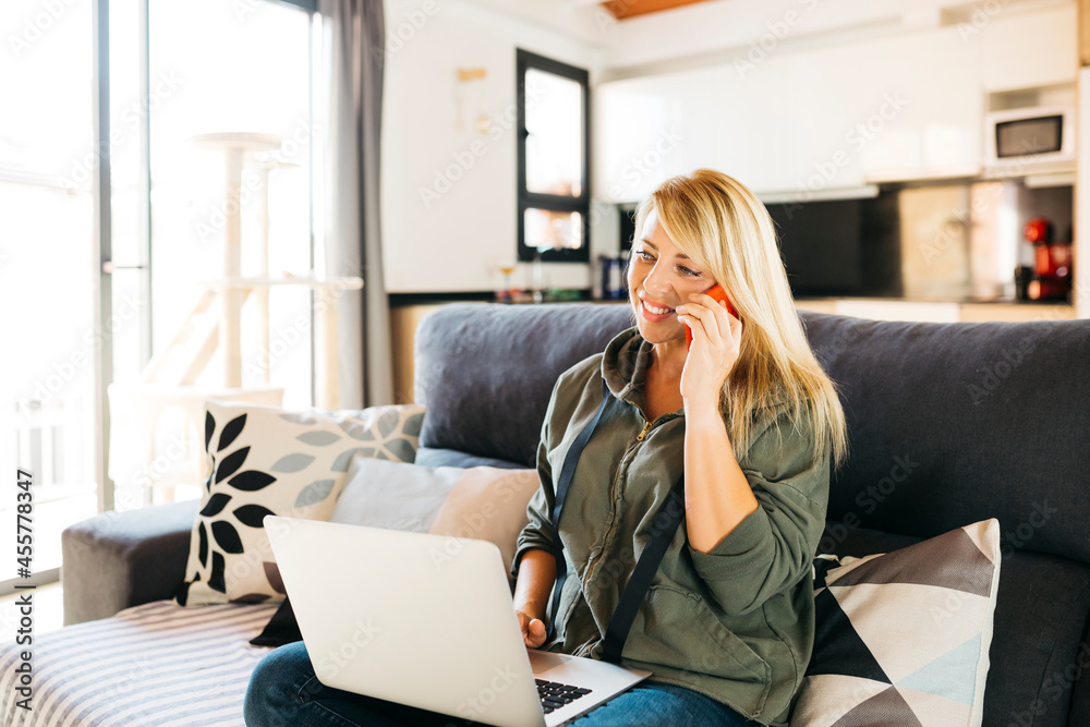 Middle age smiling blonde woman using a laptop and having a conversation on a red smartphone on the sofa of a livingroom