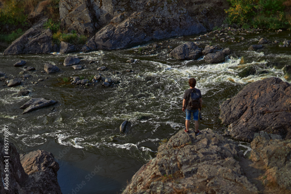 a young man with a backpack stands on the rocks by a wild river, photo from the back. The concept of travel and freedom. High quality photo