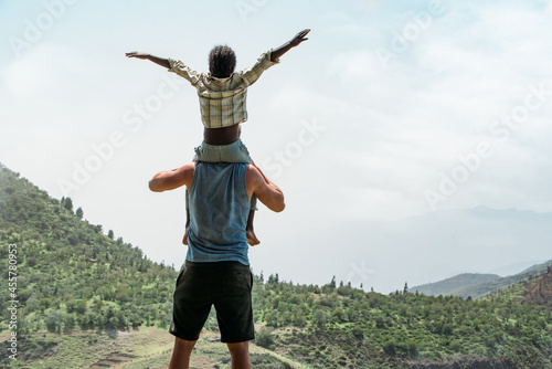 back view of caucasian father holding on shoulders black little son on top of mountain photo