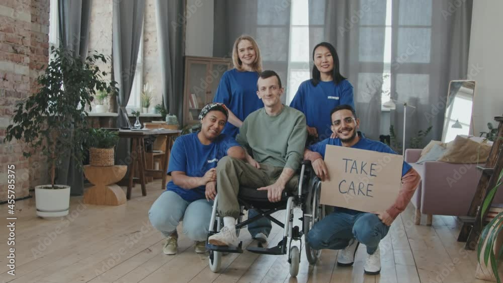 Zoom-out portrait of multi-ethnic team of volunteers in blue T-shirts and young Caucasian man with disability in wheelchair posing for camera indoors with cardboard Take Care sign Zoom-out portrait of multi-ethnic team of volunteers in blue T-shirts and young Caucasian man with disability in wheelchair posing for camera indoors with cardboard Take Care sign