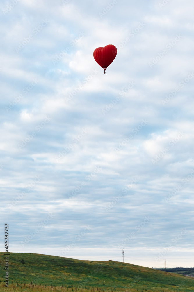 Fototapeta premium Red hot air balloon in shape of heart is landing on the green field