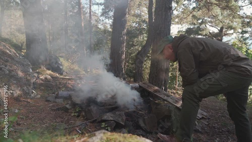 a man in a cap and hiking clothes trying to make a fire in the forest. Blowing on fire. 