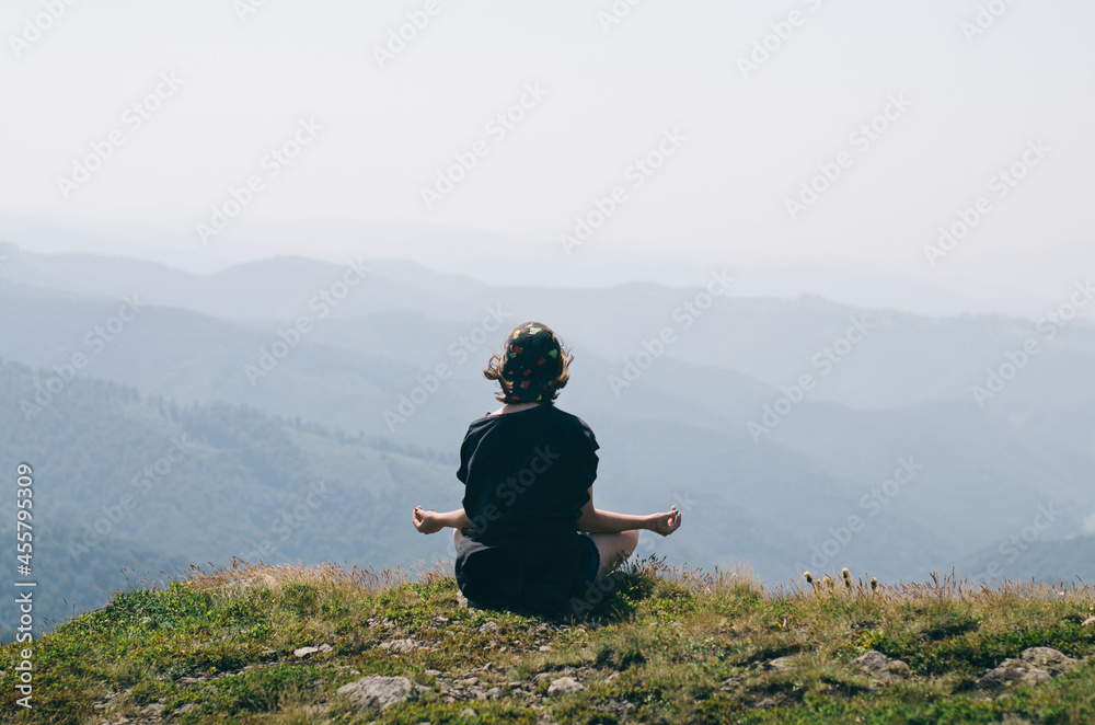 Back view of young girl sitting alone on the edge of the mountain and meditating. Female enjoying stunning landscape of mountains.