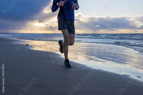Fototapeta Naklejka Na Ścianę i Meble -  Exercising outside. A jogger in lightweight blue long sleeve t-shirt, shorts and running sneakers trains to marathon, running across the Baltic Sea beach, on cloudy windy rainy early morning at down