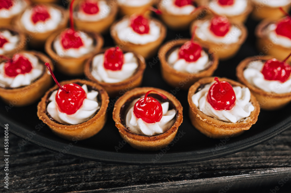 Close-up dough tartlets with cheese cream, cherries stand on a round tray, plate on a wooden table. Banquet, buffet, top view, photography.