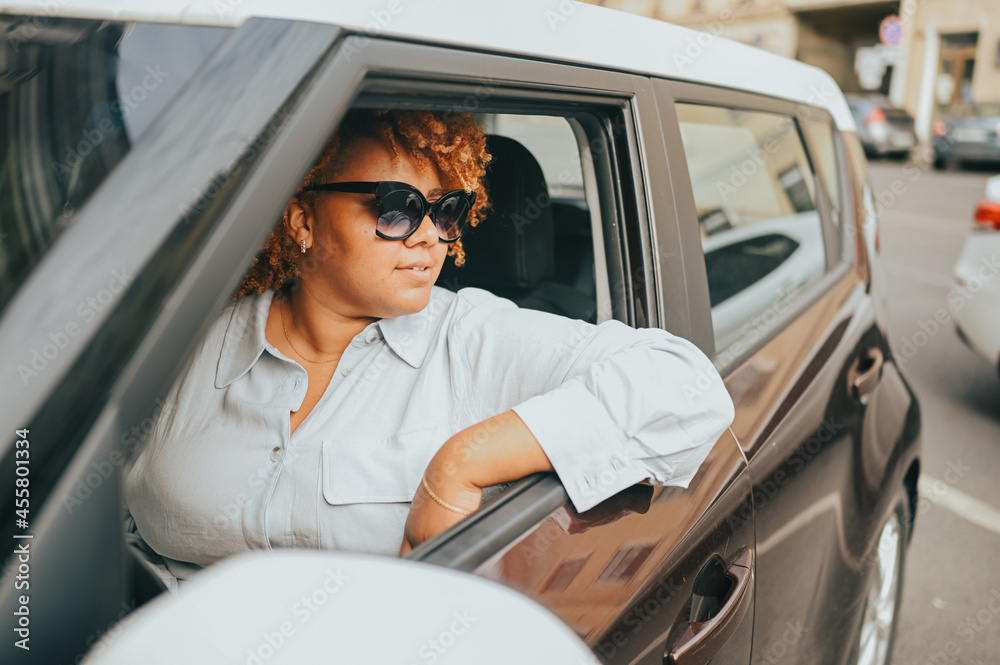 Naklejka premium Happy young smiling African American woman red afro haired driver in sun glasses sitting in new brown car, smiling looking at camera enjoying journey. Driving courses and life insurance concept.
