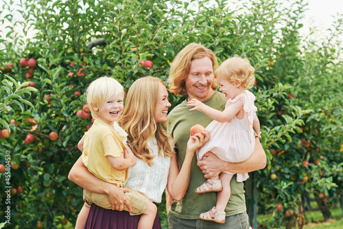 Outdoor portrait of happy young family with two little children enjoying good time in apple orchard