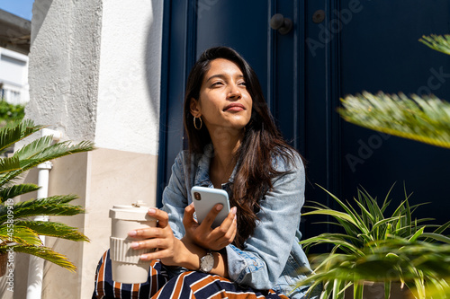 Pensive young woman holding phone and coffe cup outside