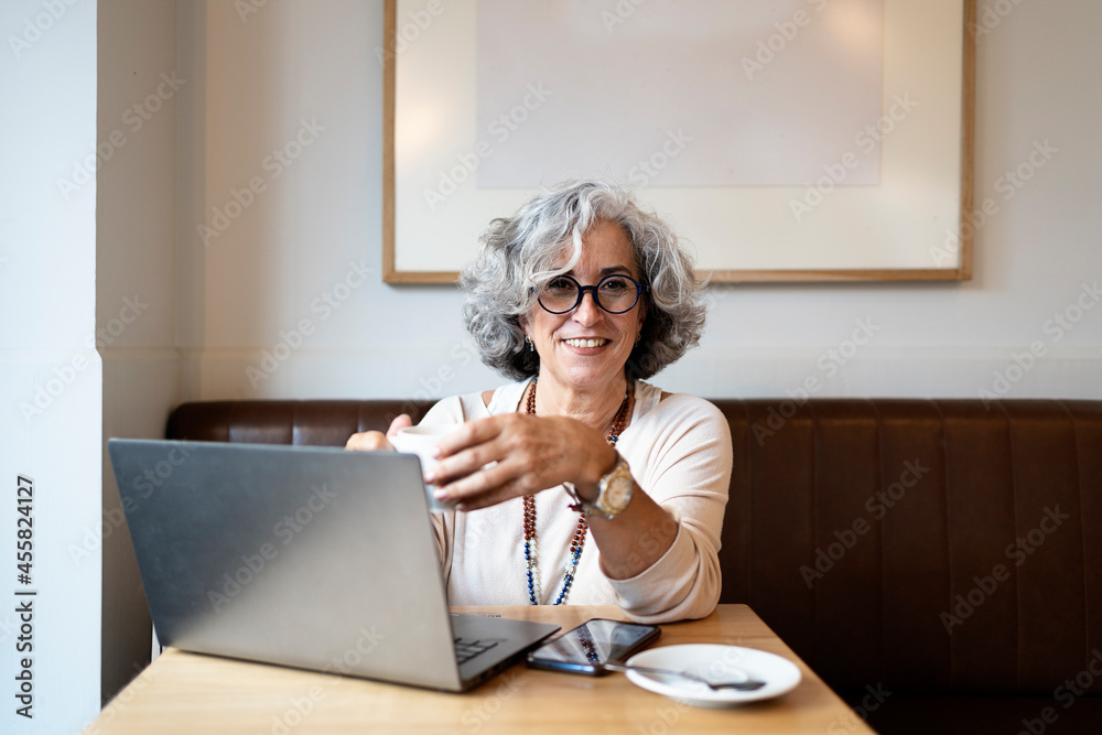 Smiling gray haired mature woman using laptop and holding cup of coffee in a cafe. Smiling gray haired mature woman using laptop and holding cup of coffee in a cafe.