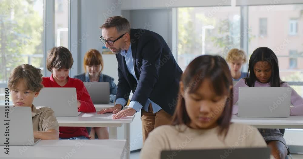 Diverse teenage students working on laptop in classroom and mature teacher help