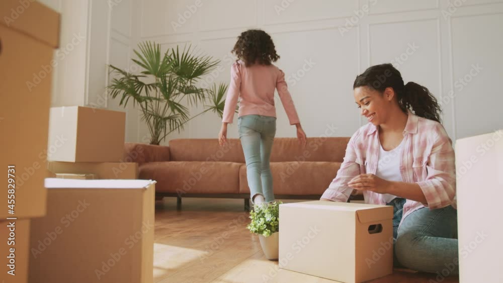Little girl helping her mom with relocation, bringing her plant and embracing with daddy on sofa, woman unpacking boxes