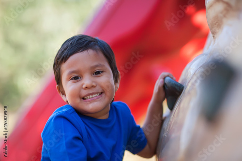4 year old boy smiles while holding on to a kids outdoor rock climbing wall.