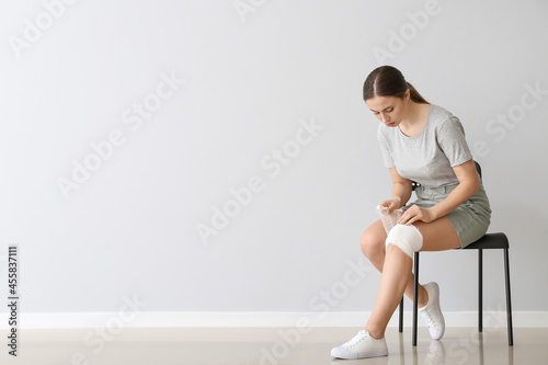 Young woman applying bandage onto her knee near light wall © Pixel-Shot