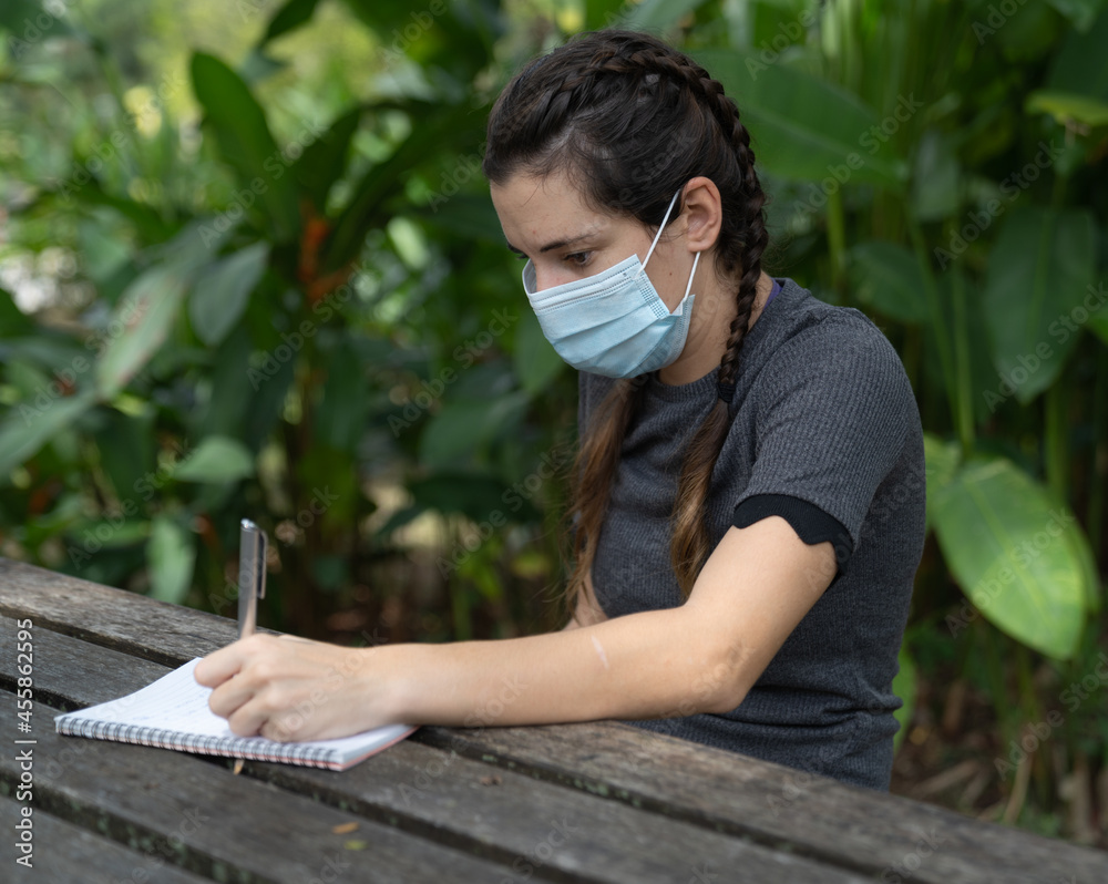 Caucasian Girl wearing a surgical mask sits in a park writing in her notepad/diary. Caucasian Girl wearing a surgical mask sits in a park writing in her notepad/diary.