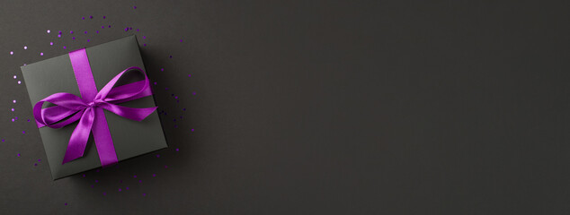 Above photo of black giftbox with purple ribbon wrapped as bow confetti and glitter around isolated on the black background