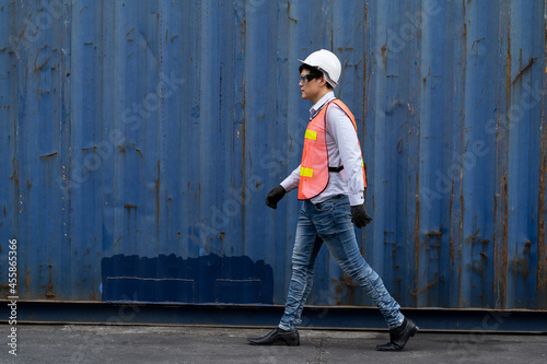 Portrait of Asian male worker or supervisor wearing safety vest, glasses and helmet at commercial dock site with container box background
