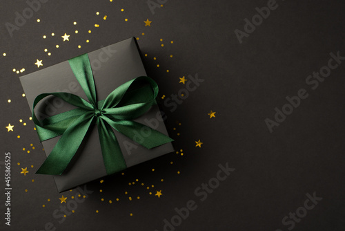 Top view photo of stylish giftbox with green ribbon bow golden stars and confetti on isolated black background with copyspace