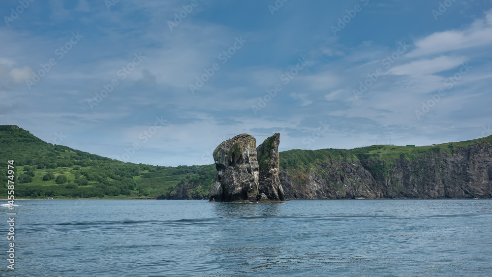High picturesque rocks rise above the surface of the Pacific Ocean. Green meadows on the coast of Kamchatka. Blue sky. Avacha Bay. Three brothers