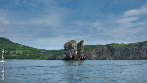 High picturesque rocks rise above the surface of the Pacific Ocean. Green meadows on the coast of Kamchatka. Blue sky. Avacha Bay. Three brothers