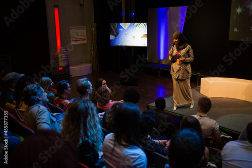 Smiling female speaker in hijab talking with microphone on stage