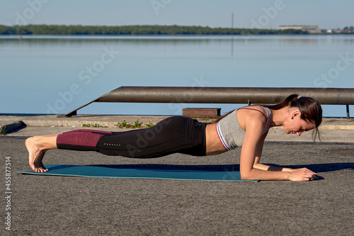 A young woman stands in a plank on the pier in the early morning, against the background of the river