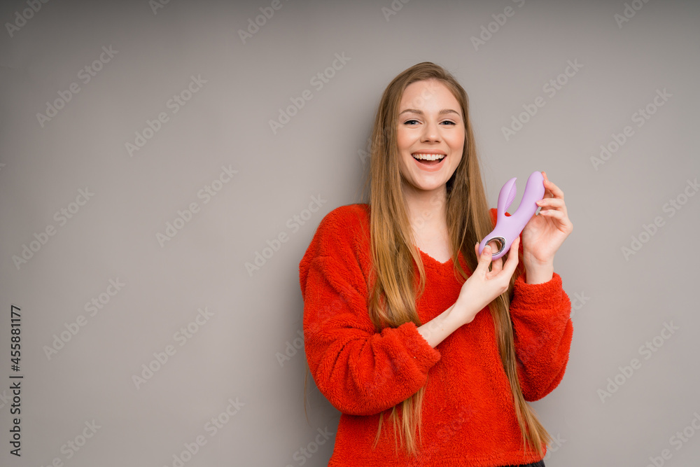 A girl with long hair smiles broadly and holds a lilac vibrator on a gray background.Useful for a sex shop A girl with long hair smiles broadly and holds a lilac vibrator on a gray background.Useful for a sex shop