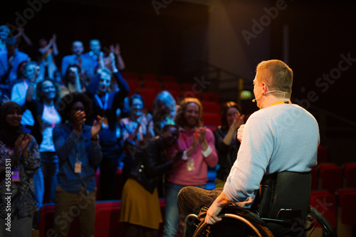 Speaker in wheelchair on stage talking to conference audience © KOTO