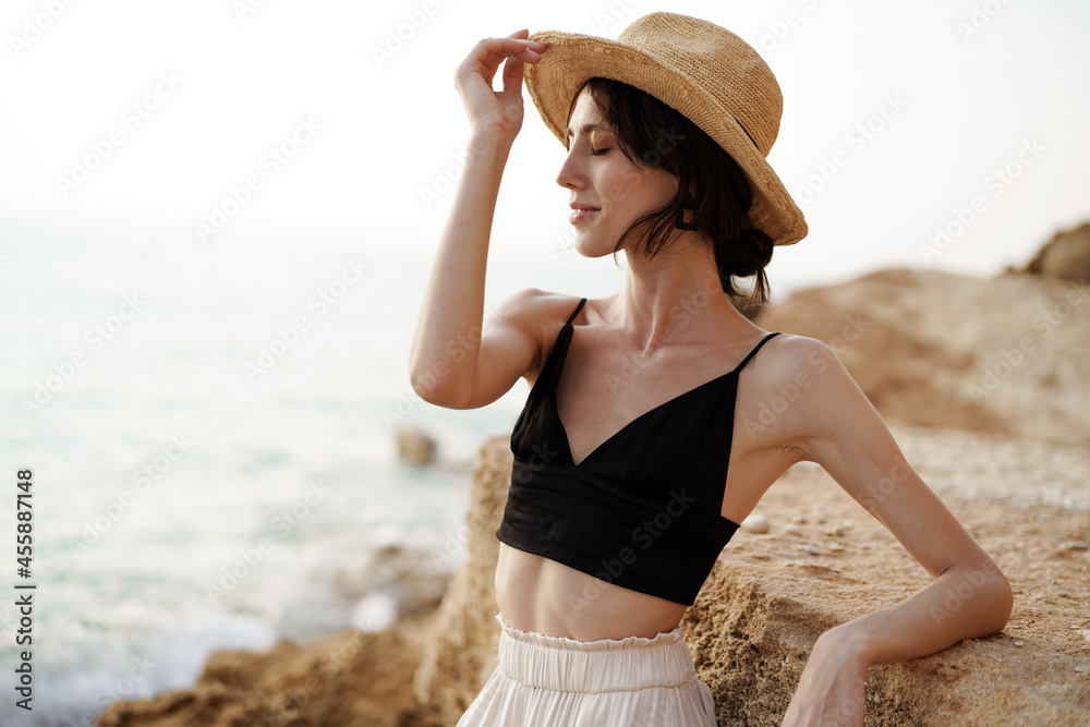 Portrait of woman in black bralette and white trousers leaning on rock at beach