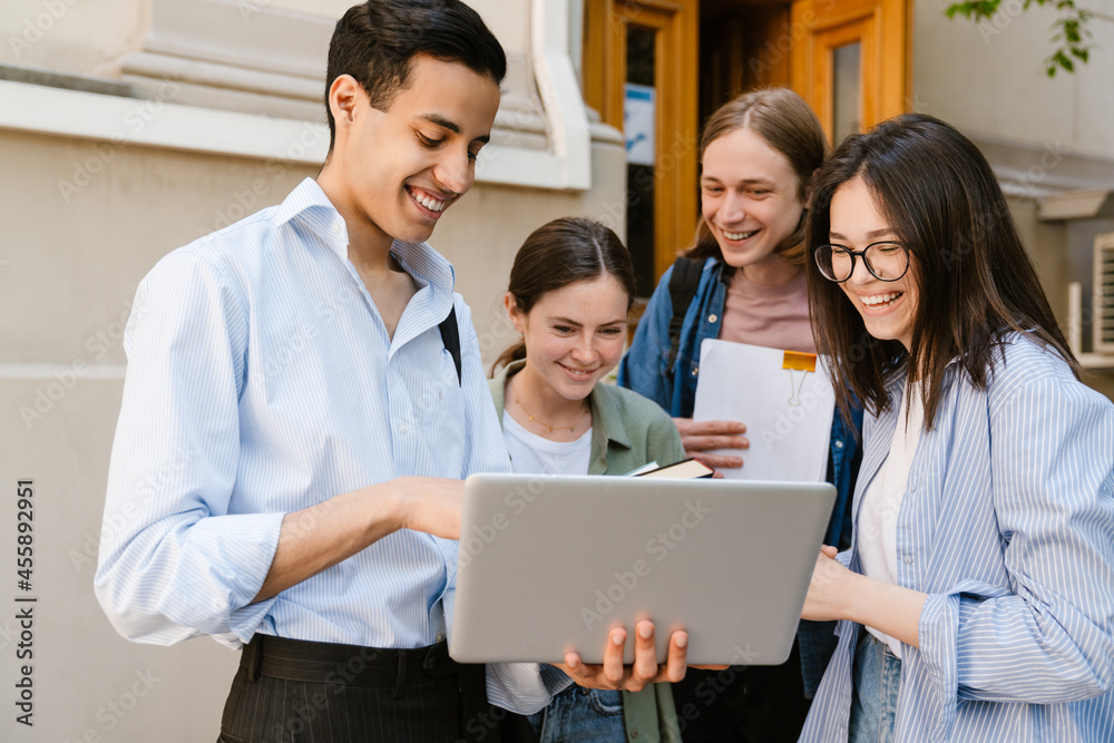 Multiracial students using laptop and laughing while standing outdoors Multiracial students using laptop and laughing while standing outdoors