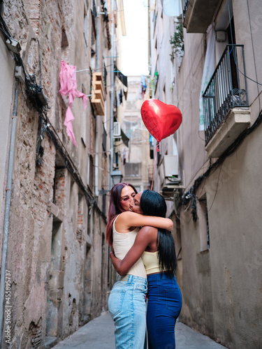 Lesbian couple hugging while holding a red heart-shaped balloon on the street.