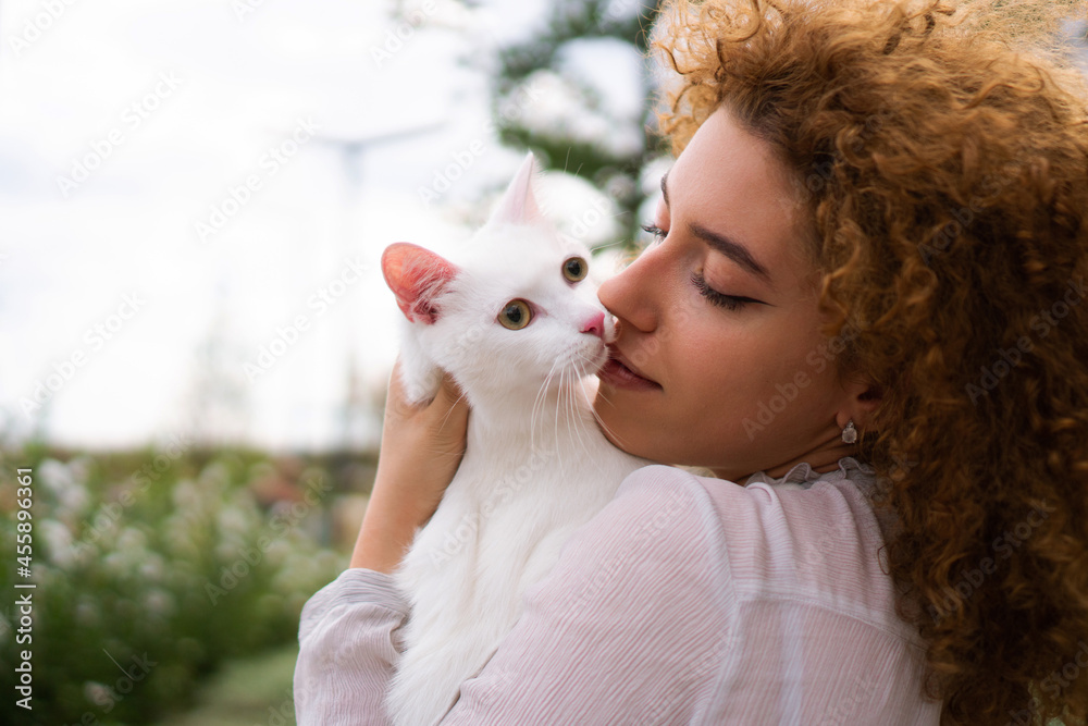 A beautiful teenage girl with curly hair holds a white cat close to her face. She is enjoying time with her pet. A beautiful teenage girl with curly hair holds a white cat close to her face. She is enjoying time with her pet.