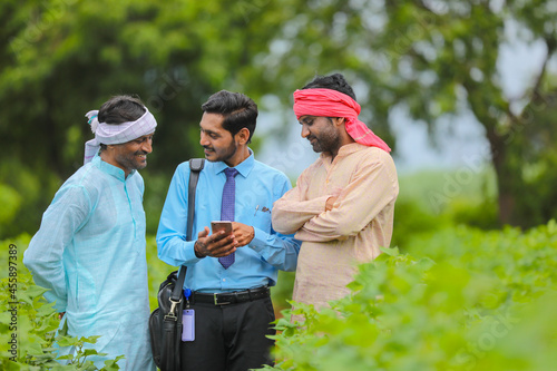 Young indian agronomist or banker showing some information to farmer in smartphone at agriculture field.