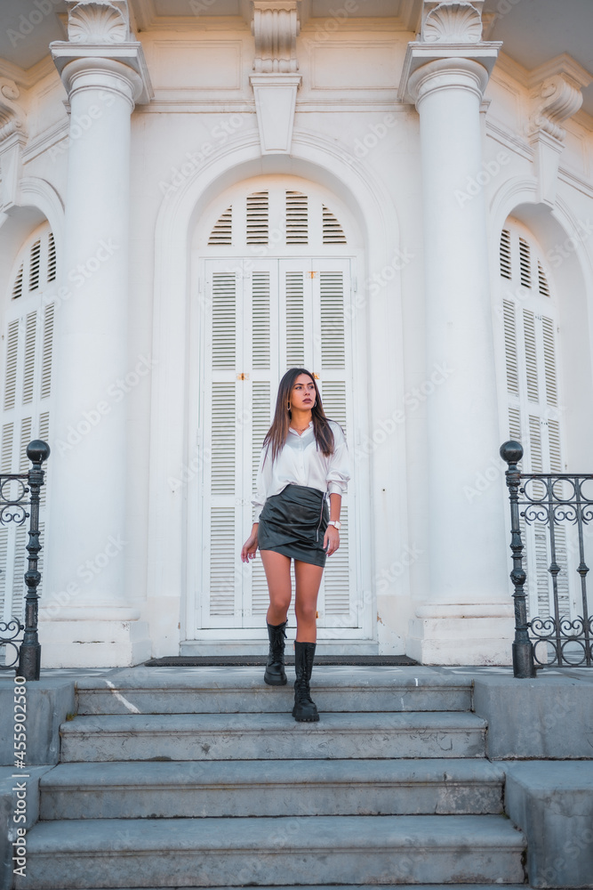 Lifestyle, fashionable pose of a young brunette with a black leather skirt and a shirt on the stairs of a white house