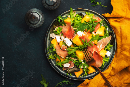 Fresh salad with cantaloupe melon, prosciutto, soft cheese and arugula on blue background, top view, copy space