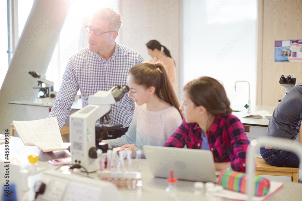 Obraz premium Male teacher helping girl students using microscope, conducting scientific experiment in laboratory classroom