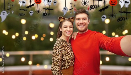 holiday and people concept - happy smiling couple in halloween costumes of devil and leopard taking selfie over roof top party background photo