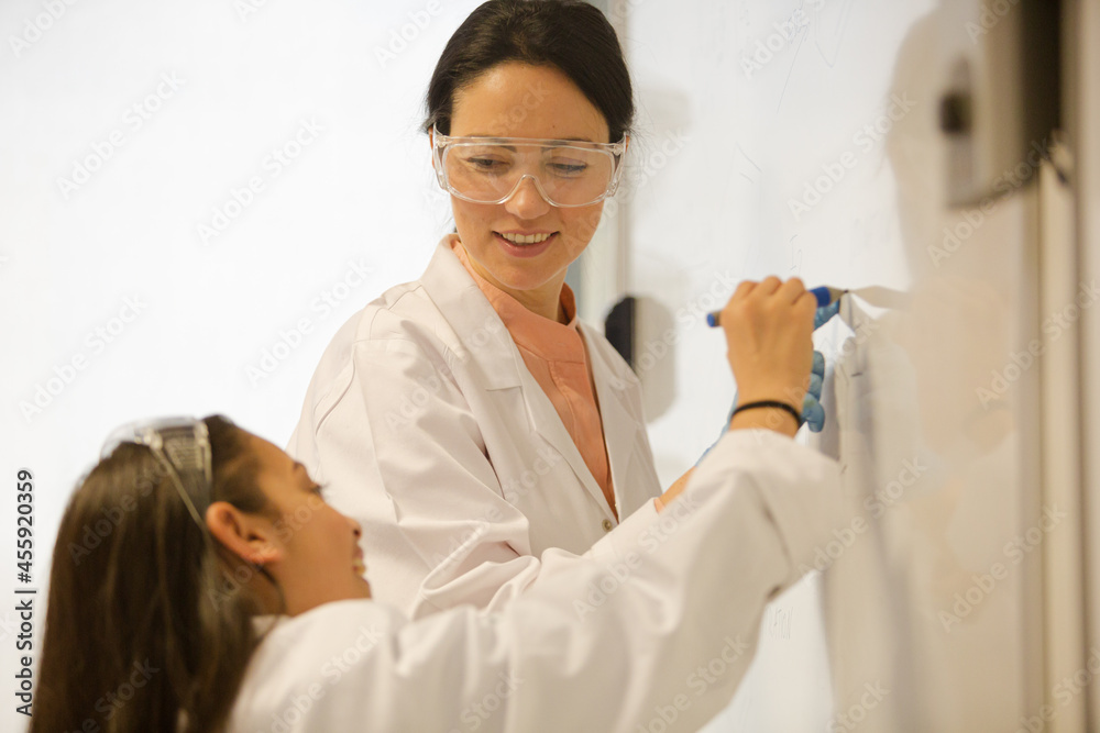 Female science teacher and girl student writing at whiteboard in classroom Female science teacher and girl student writing at whiteboard in classroom