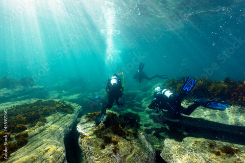 Scuba Divers Kick Over Rocks Underwater in Sun Light with Bubbles