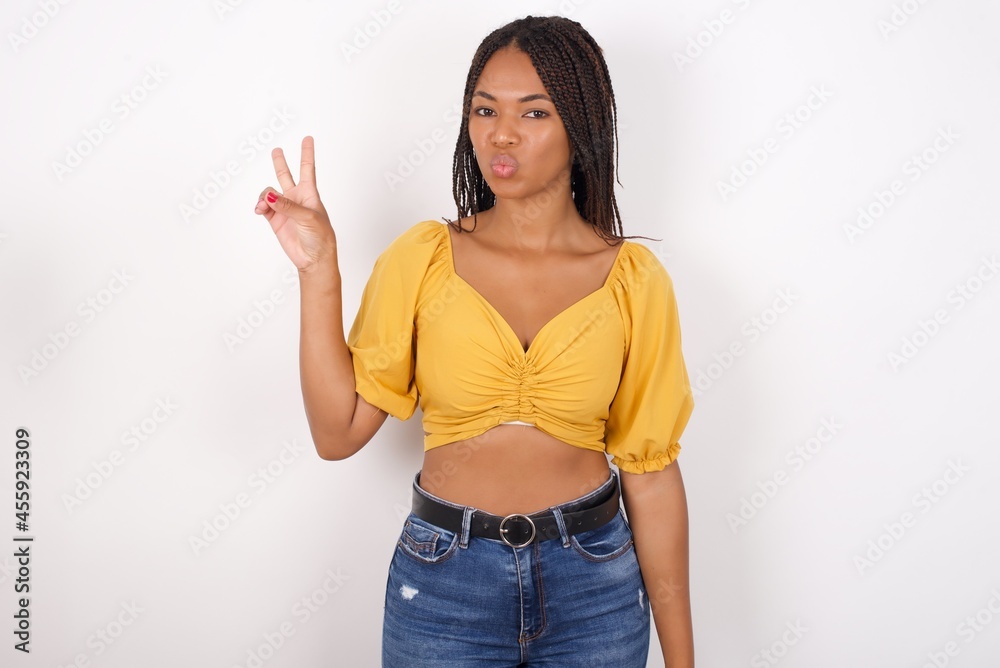 Young african american woman with braids over white wall makes peace gesture keeps lips folded shows v sign. Body language concept Young african american woman with braids over white wall makes peace gesture keeps lips folded shows v sign. Body language concept