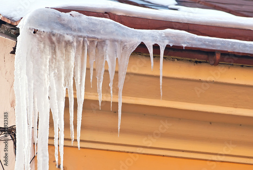 Icicles hang from the roof of the house. The concept of danger and slippery sidewalks. The beginning of spring in the city. Transparent swan icicles close up. © Ivan