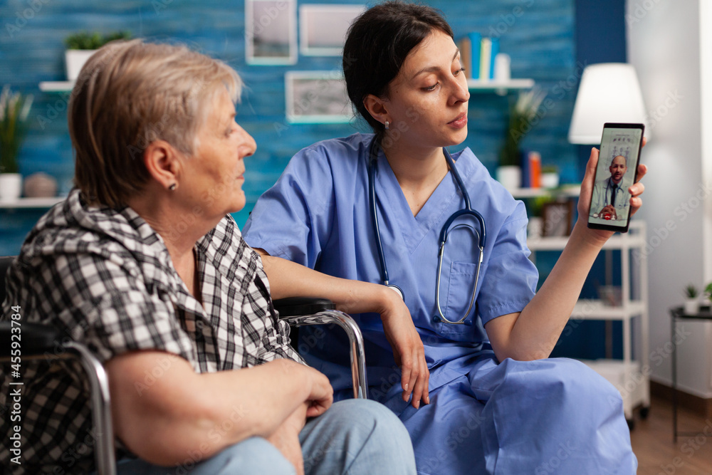 Fototapeta premium Nursing home female worker helping retired disabled elderly woman to have a video call on smartphone with her doctor. Social assistant working with people with disability in retirement home